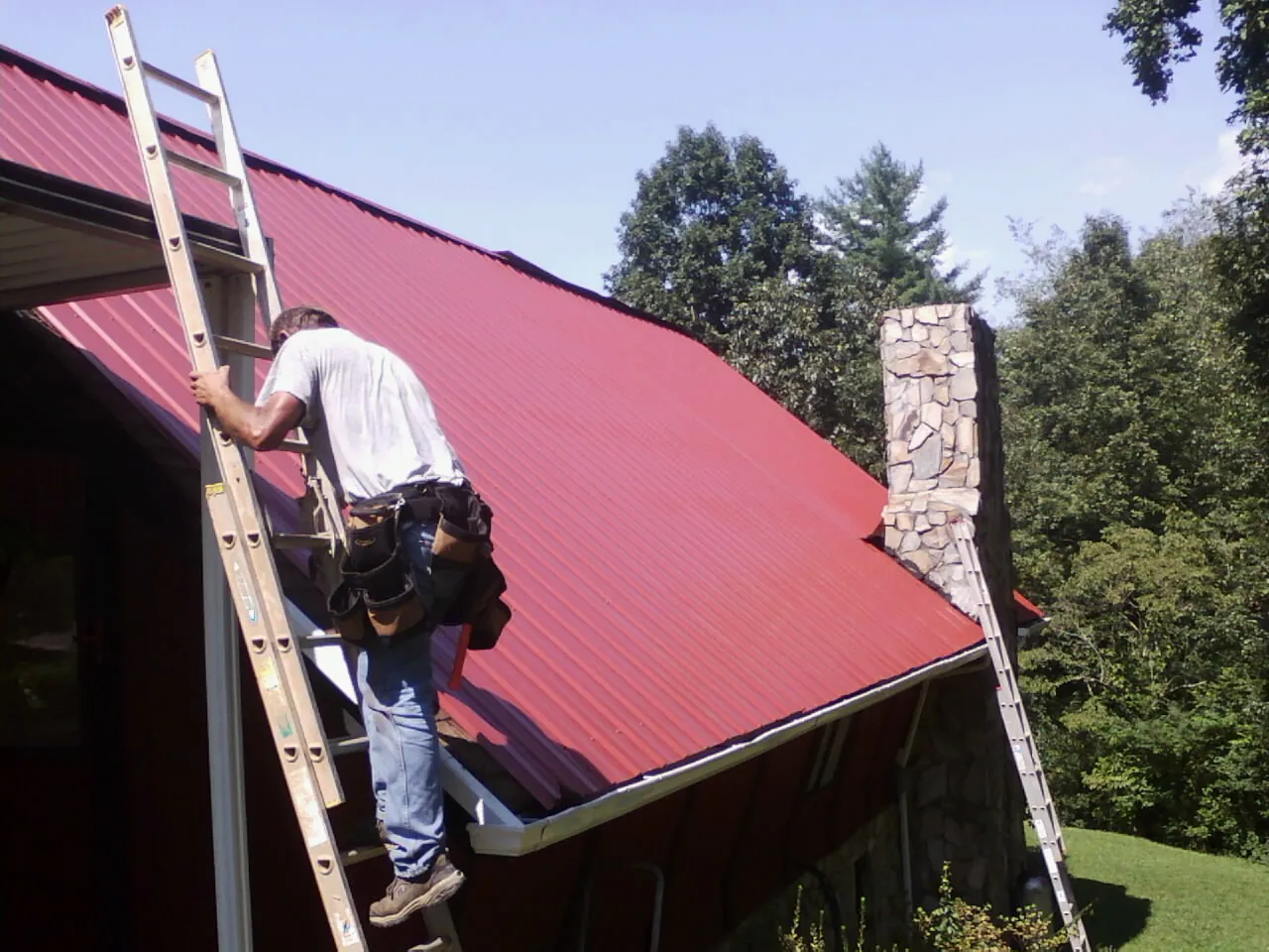 Roofer on a ladder beside a red metal roof and stone chimney — Soffit & Fascia Repair by Roofing Brothers in Fountain City